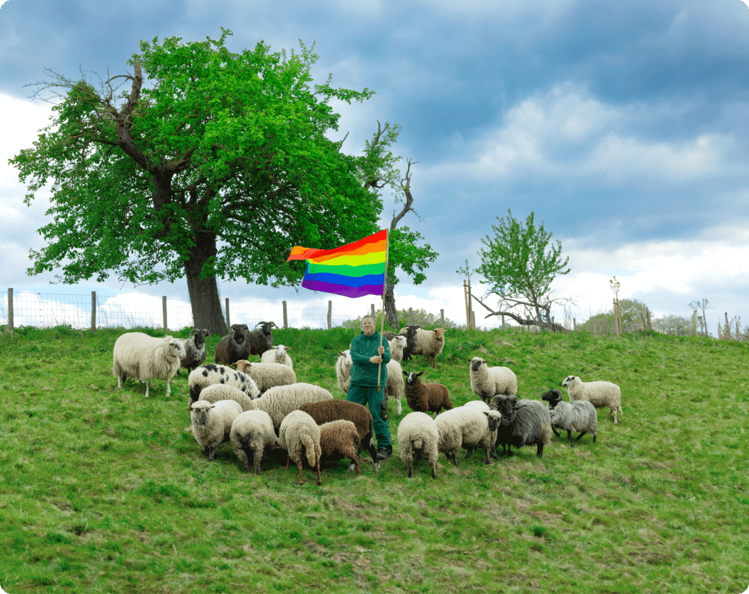 A flock of sheep on a green meadow with a shepherd holding a rainbow flag. 